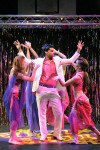a man is stood centre stage wearing a white suit and pink suit, around him are four women. they are all dancing