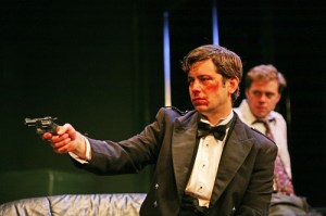 a man wearing an evening suit has blood and cuts on his face, he is pointing a hand gun at someone. A man in a shirt and tie is sat behind him watching what the gun is focused on