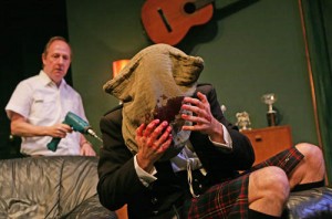 a man is wearing a Scottish kilt he has a fabric bag over his head and is holding his blood covered fingers up to a hole in the bag where his face would be. behind him is a man standing looking at him, holding a power drill in his hand.