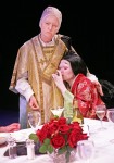 a woman wearing a traditional Chinese costume is sat down drinking a glass of wine, she is leaning on another woman standing next to her who is dressed as a priest