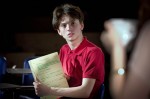 a teenage boy with brown hair wearing a red polo shirt looking out in front of him whilst holding a note book with writing in it as though he is showing someone in front of him the content
