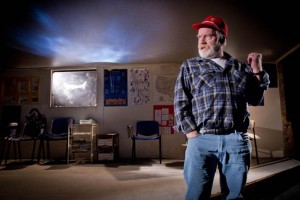 a man wearing a checkered shirt, jeans and a red hat is stood in an empty classroom with his fist in the air looking to his side. The roof of the classroom is gone and has been replaced with a night sky
