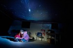 a woman is kneeling on the floor she is shielding the person next to her who is in a foetal positioned ball on the floor, with her body they are in a classroom except the ceiling is gone and has been replaced with a star filled night sky