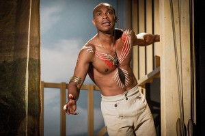 a topless man with aborigine style art painted onto his chest and wearing aboriginal style necklace and arm band, holding onto a wooden structure, looking out in front of him.