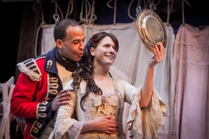 a woman in a clean, neat 1700's style dress with her hair done up nicely is standing smiling at her reflection in a silver plate which she is holding up in front of her. A man is standing behind her dressed in a smart and lavish officers uniform. The man has his hands on the woman's arms and is speaking to her.