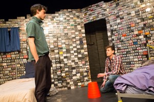 two men in a room filled with cassette tapes, one is kneeling down by a bed and overturned orange dustbin and the other standing by another bed on the opposite side looking down on the other man.