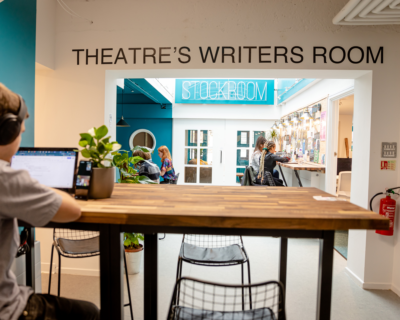 An open office space, with empty modern desks. Across the wall there is text which reads: Theatre's Writers Room